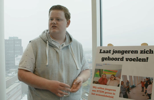 melvin presenteert en staat voor een bord met de tekst; Laat jongeren zich gehoord voelen!' en 'Wij willen jongeren een stem geven in het armoedebeleid'