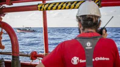 CH128955-A-Save-the-Children-team-member-looks-out-at-a-boat-crowded-with-refugees-and-migrants-in-the-Mediterranean-Sea.png