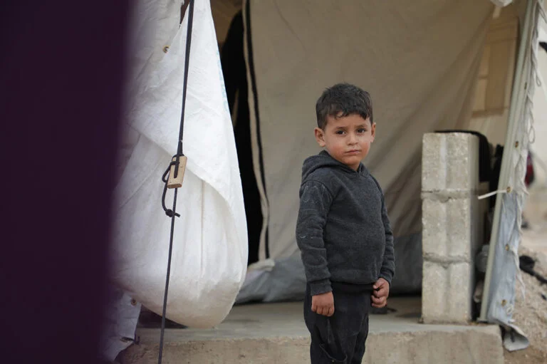 Jongetje staat voor een tent. Hij kijkt recht de camera in.