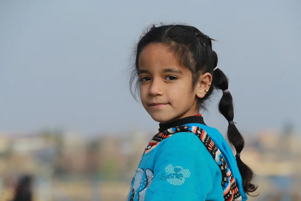 De achtjarige Amal kijkt in de camera, terwijl ze in een vluchtelingenkamp in Syrië staat