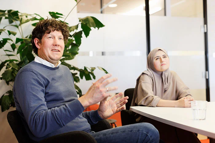 Geert Jan van Dijk zit aan een vergadertafel en praat, naast hem zit Aziza Dahman. 