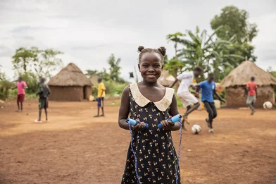 De negenjarige Amina staat in een open vlakte in haar vluchtelingenkamp in Oeganda. Ze houdt een springtouw vast, terwijl achter haar kinderen voetballen