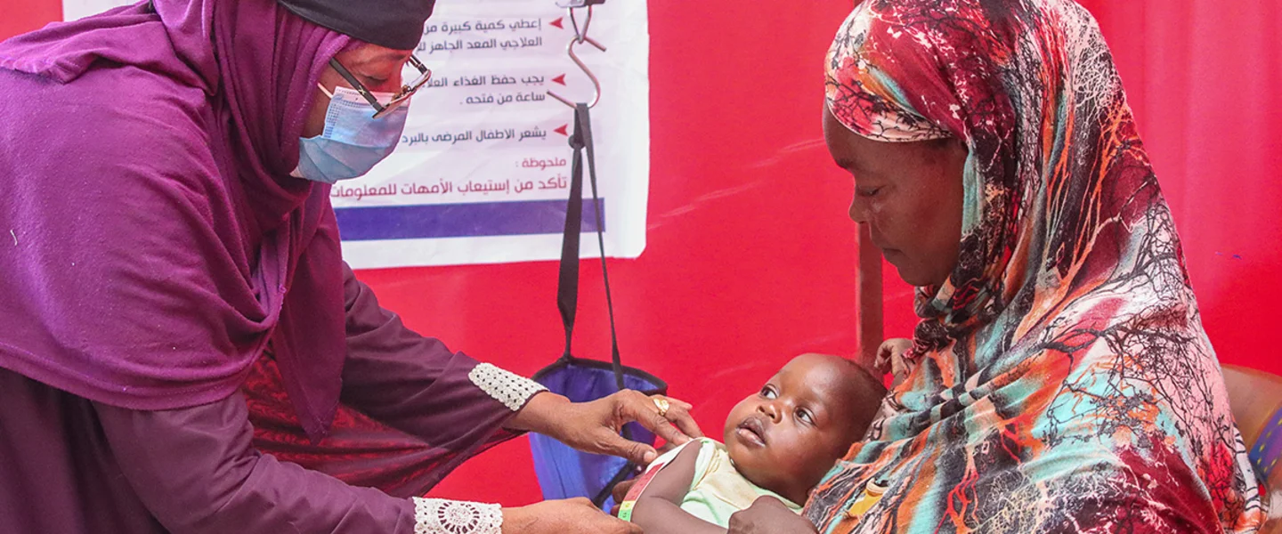 Een medewerker van Save the Children controleert of peuter Safana ondervoed is. Het meisje zit op schoot bij haar moeder.