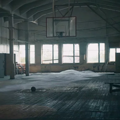 Lege gymzaal met kapotte ramen waar licht door schijnt, er ligt sneeuw op de vloer en een eenzame basketbal