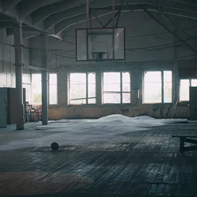 Lege gymzaal met kapotte ramen waar licht door schijnt, er ligt sneeuw op de vloer en een eenzame basketbal