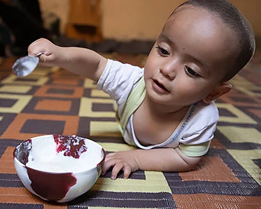Syrische dreumes ligt op de buik en staat op het punt een lepel in een bakje te steken dat voor hem/haar staat. 