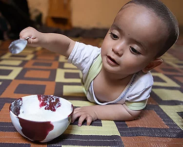 Syrische dreumes ligt op de buik en staat op het punt een lepel in een bakje te steken dat voor hem/haar staat. 
