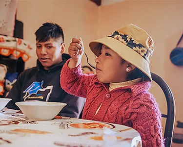 Boliviaans meisje zit aan tafel en neemt net een hap. Op de achtergrond kijkt haar broer toe.