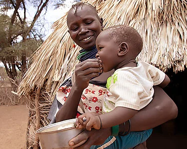 Moeder met kind op haar arm voor een rieten hut in Kenia, ze voedt haar kind eten uit een aluminium bakje dat ze vasthoud. Ze kijkt blij.