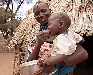 Moeder met kind op haar arm voor een rieten hut in Kenia, ze voedt haar kind eten uit een aluminium bakje dat ze vasthoud. Ze kijkt blij.