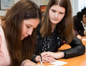 Twee mbo studenten zittend aan een tafel. Ze kijken beiden naar wat een van hen opschrijft