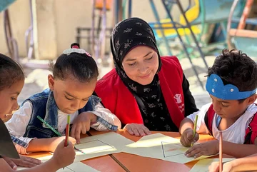 Een Save the Children medewerker in een rood hesje is aan het kleuren met kinderen in een van onze programma's